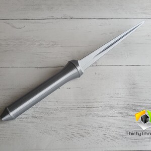May include: A silver-colored, long, thin, pointed object with a handle. The object is lying on a white wooden surface.