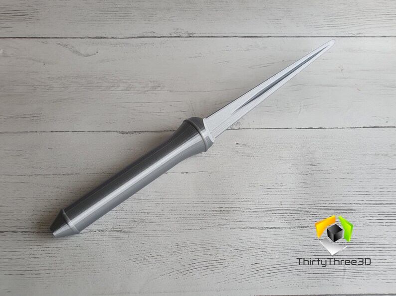 May include: A silver-colored, long, thin, pointed object with a handle. The object is lying on a white wooden surface.
