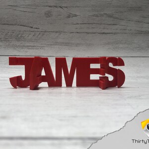 Op de afbeelding: Rood 3D-geprint naam bord met de spelling "JAMES" op een witte houten achtergrond.