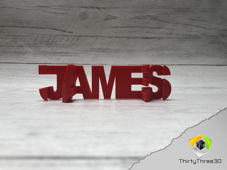 Op de afbeelding: Rood 3D-geprint naam bord met de spelling "JAMES" op een witte houten achtergrond.