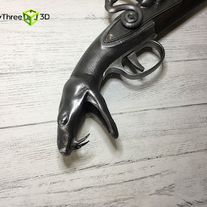 Puede incluir: R&eacute;plica gris met&aacute;lico, impresa en 3D, de una pistola de chispa. La pistola presenta un mango en forma de serpiente y un gatillo. El logotipo "ThirtyThree 3D" est&aacute; en la esquina superior izquierda.