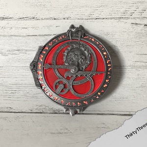 May include: A silver-colored metal pendant with a red, circular design featuring intricate details and runes. The pendant has a three-dimensional, textured surface.
