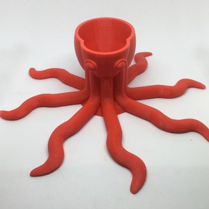 May include: A bright orange octopus-shaped ceramic holder with a cup-like top. The holder has eight tentacles.