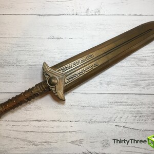 May include: A gold-colored sword with intricate details on the blade and handle. The sword is lying on a white wooden surface.
