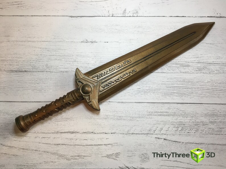 May include: A gold-colored sword with intricate details on the blade and handle. The sword is lying on a white wooden surface.