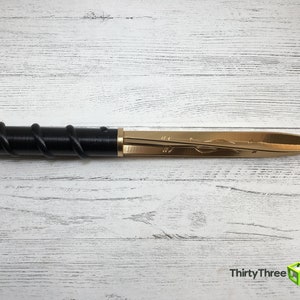 Op de afbeelding: Een decoratieve briefopener met een gouden mes en een zwarte handgreep gewikkeld met een spiraalvormig ontwerp. Het mes heeft een ge&euml;tst bloemenpatroon. De briefopener wordt gepresenteerd op een wit houten oppervlak.