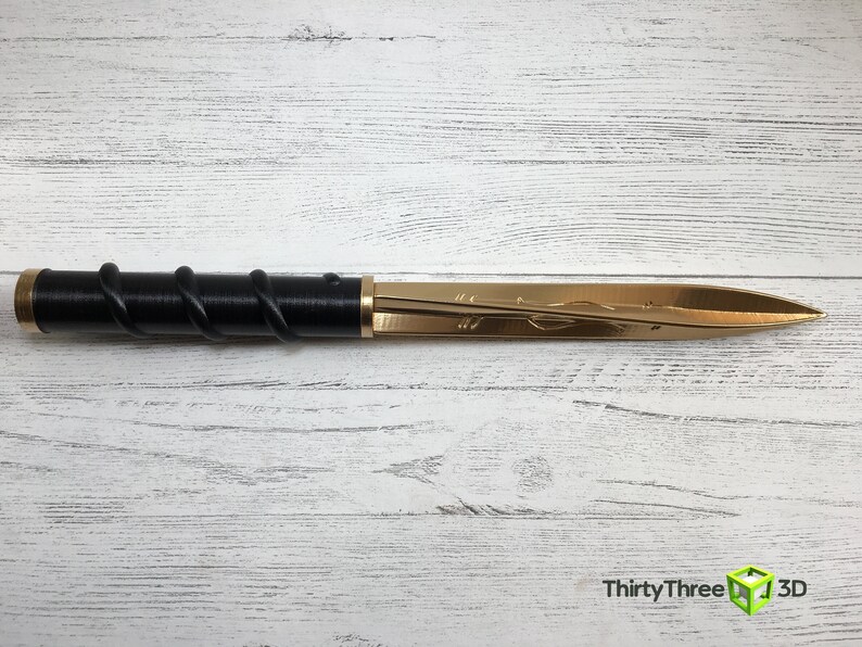 Op de afbeelding: Een decoratieve briefopener met een gouden mes en een zwarte handgreep gewikkeld met een spiraalvormig ontwerp. Het mes heeft een ge&euml;tst bloemenpatroon. De briefopener wordt gepresenteerd op een wit houten oppervlak.