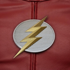 Puede incluir: Primer plano de una prenda de cuero roja con un emblema dorado y plateado. El emblema presenta un dise&ntilde;o de rayo dorado sobre un fondo circular plateado. El cuero tiene un aspecto texturizado.