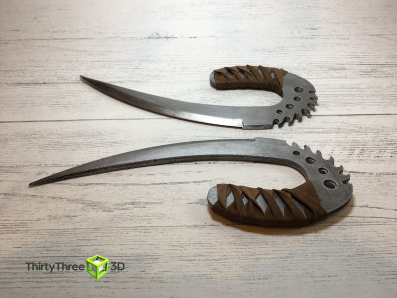 May include: Two silver-colored, curved, and serrated knives with brown leather handles. The knives are on a white wooden surface.
