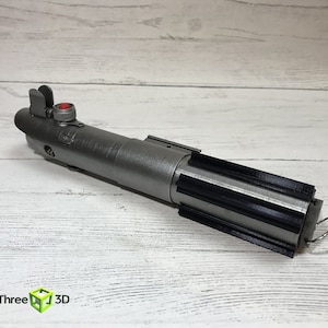 May include: A silver lightsaber prop with a black grip and a red button. The lightsaber is on a white wooden surface.