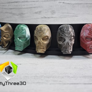 May include: A set of seven 3D printed masks in various colors, including blue, gold, green, brown, silver, and red. The masks are displayed on a black stand. The masks have a skull-like design with closed eyes and a raised nose.