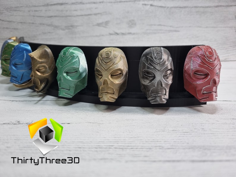 May include: A set of seven 3D printed masks in various colors, including blue, gold, green, brown, silver, and red. The masks are displayed on a black stand. The masks have a skull-like design with closed eyes and a raised nose.