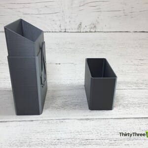 May include: Two gray 3D printed boxes, one tall and narrow, the other short and wide. The tall box has a design on the front.