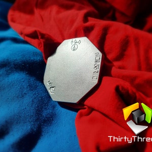 Può includere: Un oggetto metallico ottagonale argentato con il testo "3D Printed" e "ThirtyThree3D" su di esso. L'oggetto è su un tessuto rosso e blu.