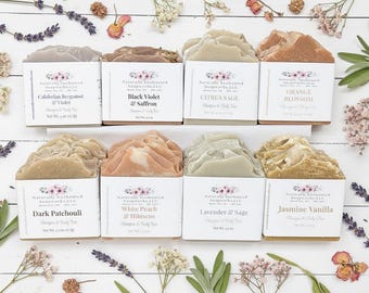 May include: An overhead shot of eight bars of soap, each in a white box with a label. The soaps are arranged on a white wooden surface, surrounded by dried flowers and sprigs of lavender. The labels read: Calabrian Bergamot & Violet, Black Violet & Saffron, Citrus Sage, Orange Blossom, Dark Patchouli, White Peach & Hibiscus, Lavender & Sage, and Jasmine Vanilla.