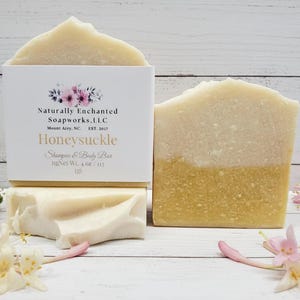 K&ouml;nnte beinhalten: Zweifarbige Honeysuckle Shampoo- und K&ouml;rperseifen mit einer wei&szlig;en Schachtel mit floralen Akzenten und Text. Die Seifen wiegen jeweils 113 g. Rosa und wei&szlig;e Blumen sind um die Seifen auf einer wei&szlig;en Holzoberfl&auml;che verstreut.