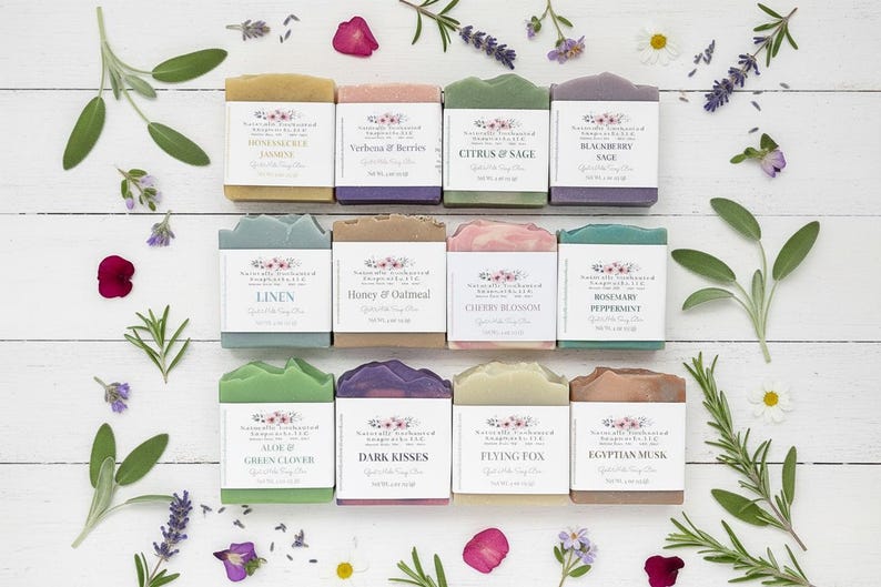 May include: Twelve rectangular bars of soap arranged in three rows, each wrapped in a white paper label with floral branding and text including Honeysuckle Jasmine, Verbena & Berries, Citrus & Sage, Blackberry Sage, Linen, Honey & Oatmeal, Cherry Blossom, Rosemary Peppermint, Aloe & Green Clover, Dark Kisses, Flying Fox, and Egyptian Musk.