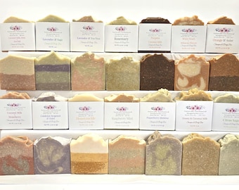 Op de afbeelding: Een collectie van eenentwintig rechthoekige stukken zeep, gerangschikt in drie rijen op een getrapte display. Elk stuk heeft een unieke kleur, textuur en een label van Naturally Enchanted Soapworks LLC met een gewicht van 113 gram.