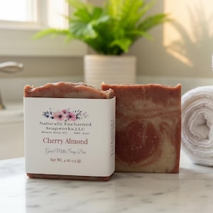 May include: A rectangular bar of marbled reddish-brown and cream-colored soap wrapped in a white paper label. The label features floral graphics and text reading Naturally Enchanted Soapworks, LLC, Cherry Almond, Goat Milk Soap Bar, Net Wt. 4 oz.