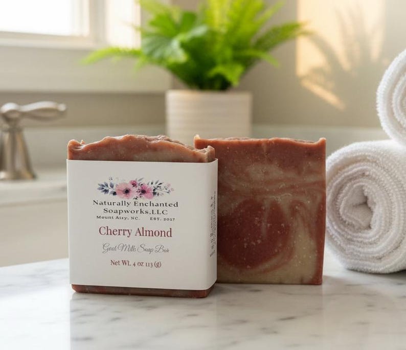 May include: A rectangular bar of marbled reddish-brown and cream-colored soap wrapped in a white paper label. The label features floral graphics and text reading Naturally Enchanted Soapworks, LLC, Cherry Almond, Goat Milk Soap Bar, Net Wt. 4 oz.