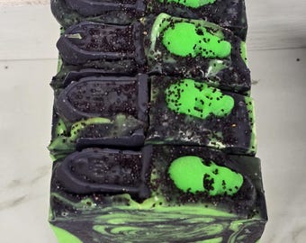 May include: A rectangular bar of soap featuring a black and green marbled base. The top is decorated with a dark gray tombstone-shaped topper and a bright neon green skull accent, all dusted with fine black glitter particles across the surface.