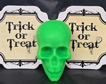 May include: A bright neon green skull figurine positioned in front of two identical square-shaped decorative signs. Each sign features a cream-colored background with a black ornate border and the black text Trick or Treat printed in a stylized font.