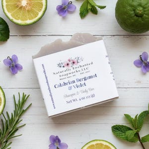 K&ouml;nnte beinhalten: Ein Seifenst&uuml;ck mit der Aufschrift "Calabrian Bergamot & Violet" ist umgeben von geschnittenen Zitrusfr&uuml;chten, Rosmarinzweigen, Minzbl&auml;ttern und kleinen lila Blumen. Die Seife wiegt 113 g.