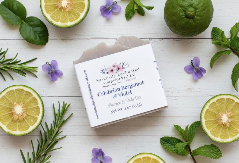 K&ouml;nnte beinhalten: Ein Seifenst&uuml;ck mit der Aufschrift "Calabrian Bergamot & Violet" ist umgeben von geschnittenen Zitrusfr&uuml;chten, Rosmarinzweigen, Minzbl&auml;ttern und kleinen lila Blumen. Die Seife wiegt 113 g.