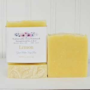 May include: Two rectangular bars of yellow goat milk soap. One bar is wrapped in a white paper label featuring a floral graphic, the text Naturally Enchanted Soapworks, LLC, Mount Airy, NC, Est. 2017, Lemon, Goat Milk Soap Bar, and Net Wt. 4 oz.
