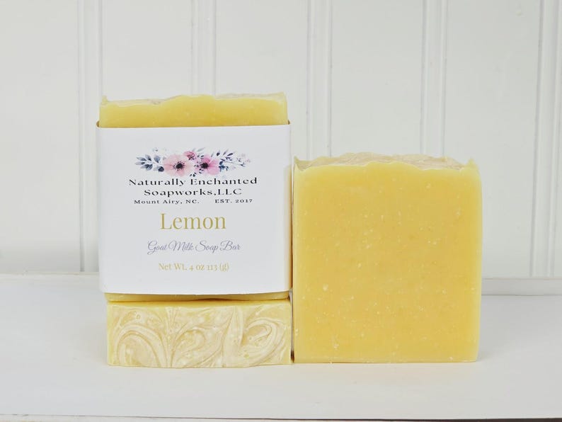 May include: Two rectangular bars of yellow goat milk soap. One bar is wrapped in a white paper label featuring a floral graphic, the text Naturally Enchanted Soapworks, LLC, Mount Airy, NC, Est. 2017, Lemon, Goat Milk Soap Bar, and Net Wt. 4 oz.