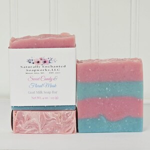 May include: A rectangular goat milk soap bar with horizontal stripes of pink and light blue. A white paper label wraps around the soap, featuring the text Naturally Enchanted Soapworks, LLC, Sweet Candy & Floral Musk, and Net Wt. 4 oz.