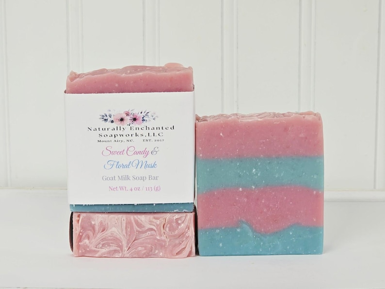 May include: A rectangular goat milk soap bar with horizontal stripes of pink and light blue. A white paper label wraps around the soap, featuring the text Naturally Enchanted Soapworks, LLC, Sweet Candy & Floral Musk, and Net Wt. 4 oz.