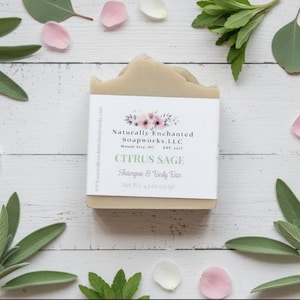 K&ouml;nnte beinhalten: Ein hellbraunes Citrus Sage Shampoo- und K&ouml;rperseifenst&uuml;ck mit einem wei&szlig;en Etikett mit floralen Akzenten und Text. Die Seife ist von gr&uuml;nen Bl&auml;ttern, rosa und wei&szlig;en Bl&uuml;tenbl&auml;ttern umgeben. Die Seife wiegt 128 Gramm.