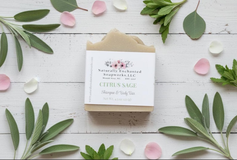 K&ouml;nnte beinhalten: Ein hellbraunes Citrus Sage Shampoo- und K&ouml;rperseifenst&uuml;ck mit einem wei&szlig;en Etikett mit floralen Akzenten und Text. Die Seife ist von gr&uuml;nen Bl&auml;ttern, rosa und wei&szlig;en Bl&uuml;tenbl&auml;ttern umgeben. Die Seife wiegt 128 Gramm.