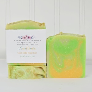 May include: Two bars of goat milk soap with a marbled lime green and orange swirl pattern. One bar is wrapped in a white paper label featuring the text Naturally Enchanted Soapworks, LLC, Mount Airy, NC, Est. 2017, Sour Candies, Goat Milk Soap Bar, Net Wt. 4 oz.