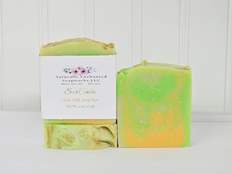 May include: Two bars of goat milk soap with a marbled lime green and orange swirl pattern. One bar is wrapped in a white paper label featuring the text Naturally Enchanted Soapworks, LLC, Mount Airy, NC, Est. 2017, Sour Candies, Goat Milk Soap Bar, Net Wt. 4 oz.