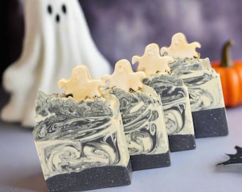 May include: Four bars of handmade soap with a black and white swirl design, topped with a small ghost-shaped decoration. A white ghost figure and an orange pumpkin are in the background, suggesting a Halloween theme.