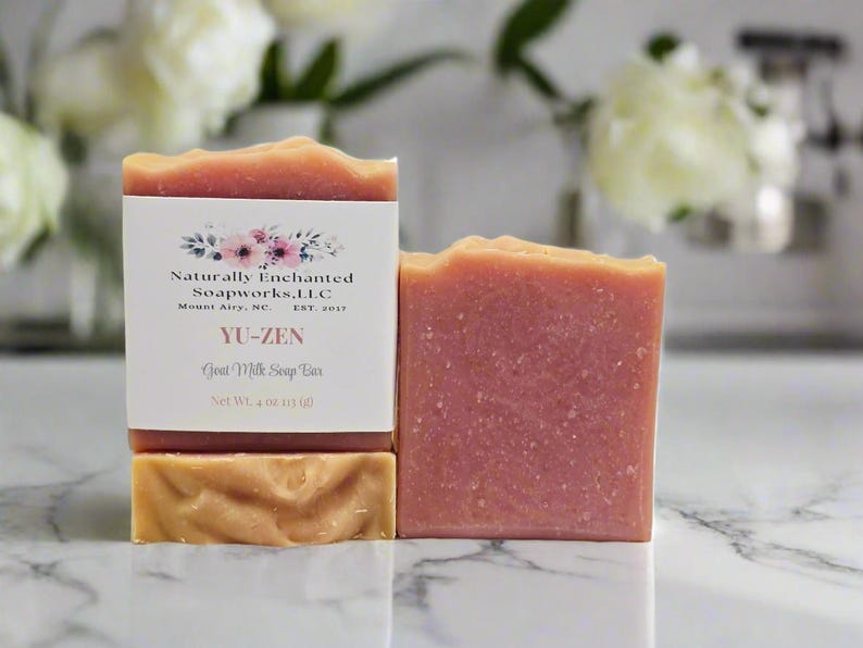 May include: A rectangular bar of pink goat milk soap with a textured top, wrapped in a white paper label featuring floral graphics and the text Naturally Enchanted Soapworks, L.L.C, Mount Airy, NC, EST. 2017, YU-ZEN, Goat Milk Soap Bar, Net Wt. 4 oz.