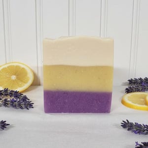 May include: A rectangular bar of soap featuring three distinct horizontal layers of color: a creamy off-white top, a vibrant yellow middle, and a deep purple base. The soap is surrounded by fresh sprigs of purple lavender and bright yellow lemon slices.