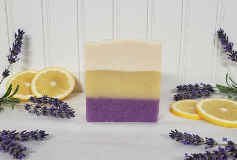 May include: A rectangular bar of soap featuring three distinct horizontal layers of color: a creamy off-white top, a vibrant yellow middle, and a deep purple base. The soap is surrounded by fresh sprigs of purple lavender and bright yellow lemon slices.