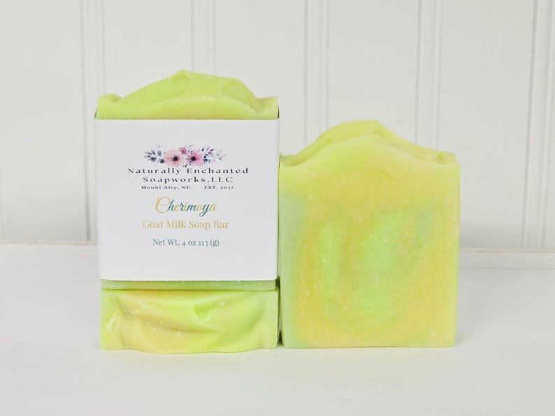 May include: Two bars of light yellow and green goat milk soap. One bar is wrapped in a white paper label with the text Naturally Enchanted Soapworks, LLC, Mount Airy, NC, Est 2017, Cherimoya, Goat Milk Soap Bar, Net Wt. 4 oz 113 g.