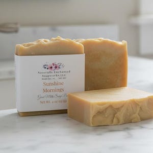 May include: Two rectangular bars of light tan goat milk soap. One bar is wrapped in a white paper label with a floral graphic and text reading Naturally Enchanted Soapworks, LLC, Sunshine Mornings, Goat Milk Soap Bar, Net Wt. 4 oz. 113 (g).