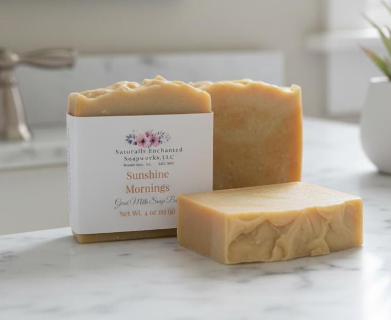 May include: Two rectangular bars of light tan goat milk soap. One bar is wrapped in a white paper label with a floral graphic and text reading Naturally Enchanted Soapworks, LLC, Sunshine Mornings, Goat Milk Soap Bar, Net Wt. 4 oz. 113 (g).