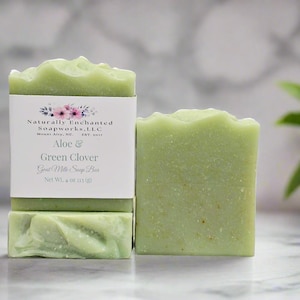 May include: Two rectangular bars of light green goat milk soap. One bar features a white paper label with floral graphics and text reading Naturally Enchanted Soapworks, L.L.C, Mount Airy, NC, EST. 2017, Aloe & Green Clover, Goat Milk Soap Bar, Net Wt. 4 oz (113 g).