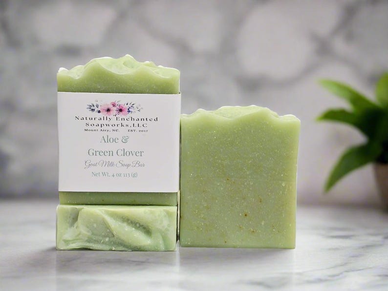 May include: Two rectangular bars of light green goat milk soap. One bar features a white paper label with floral graphics and text reading Naturally Enchanted Soapworks, L.L.C, Mount Airy, NC, EST. 2017, Aloe & Green Clover, Goat Milk Soap Bar, Net Wt. 4 oz (113 g).