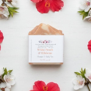 K&ouml;nnte beinhalten: Ein Seifenst&uuml;ck mit einem wei&szlig;en Etikett mit der Aufschrift "White Peach & Hibiscus Shampoo & Body Bar, Net Wt. 119g" ist von roten Hibiskusbl&uuml;ten und wei&szlig;en Bl&uuml;ten auf einer wei&szlig;en Oberfl&auml;che umgeben. Die Seife hat eine hellpfirsichfarbene Farbe.