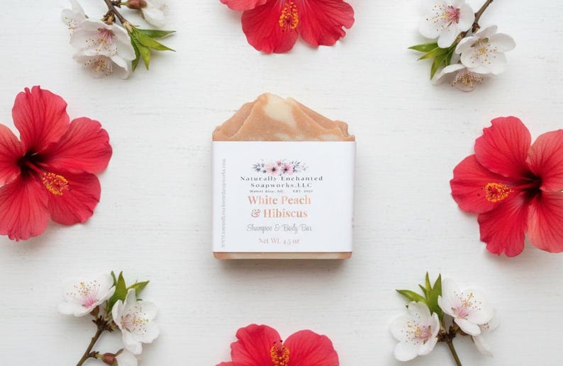 K&ouml;nnte beinhalten: Ein Seifenst&uuml;ck mit einem wei&szlig;en Etikett mit der Aufschrift "White Peach & Hibiscus Shampoo & Body Bar, Net Wt. 119g" ist von roten Hibiskusbl&uuml;ten und wei&szlig;en Bl&uuml;ten auf einer wei&szlig;en Oberfl&auml;che umgeben. Die Seife hat eine hellpfirsichfarbene Farbe.