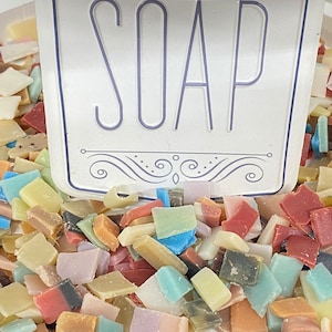 May include: A close-up of a bowl filled with colorful soap scraps. The scraps are in various shapes and sizes, and some are arranged in a pattern. A white sign with the word "SOAP" in purple lettering is partially visible in the top center of the image.