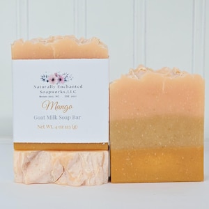 May include: Two rectangular bars of goat milk soap. One bar is wrapped in a white paper label with floral graphics and text reading Naturally Enchanted Soapworks, LLC, Mango, Goat Milk Soap Bar, Net Wt. 4 oz 113 g. The soap has layered orange hues.