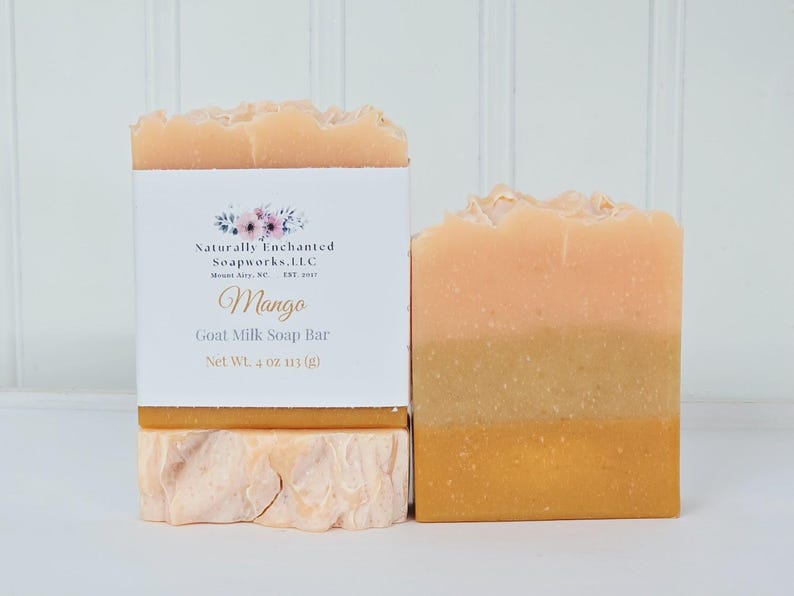 May include: Two rectangular bars of goat milk soap. One bar is wrapped in a white paper label with floral graphics and text reading Naturally Enchanted Soapworks, LLC, Mango, Goat Milk Soap Bar, Net Wt. 4 oz 113 g. The soap has layered orange hues.
